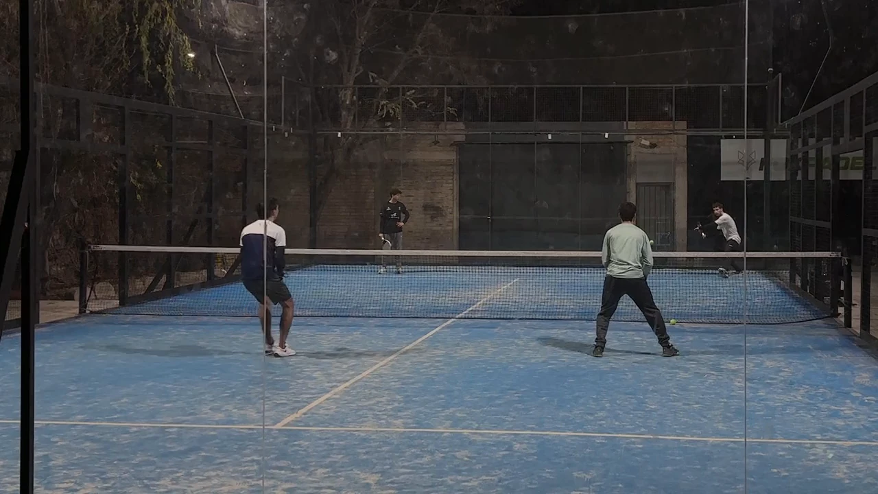 Cuatro jugadores de pádel en una cancha azul, filmados desde detrás del vidrio de fondo.